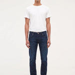 A man wearing dark blue slim-fit jeans, a white t-shirt, and black shoes, standing against a plain white studio background.