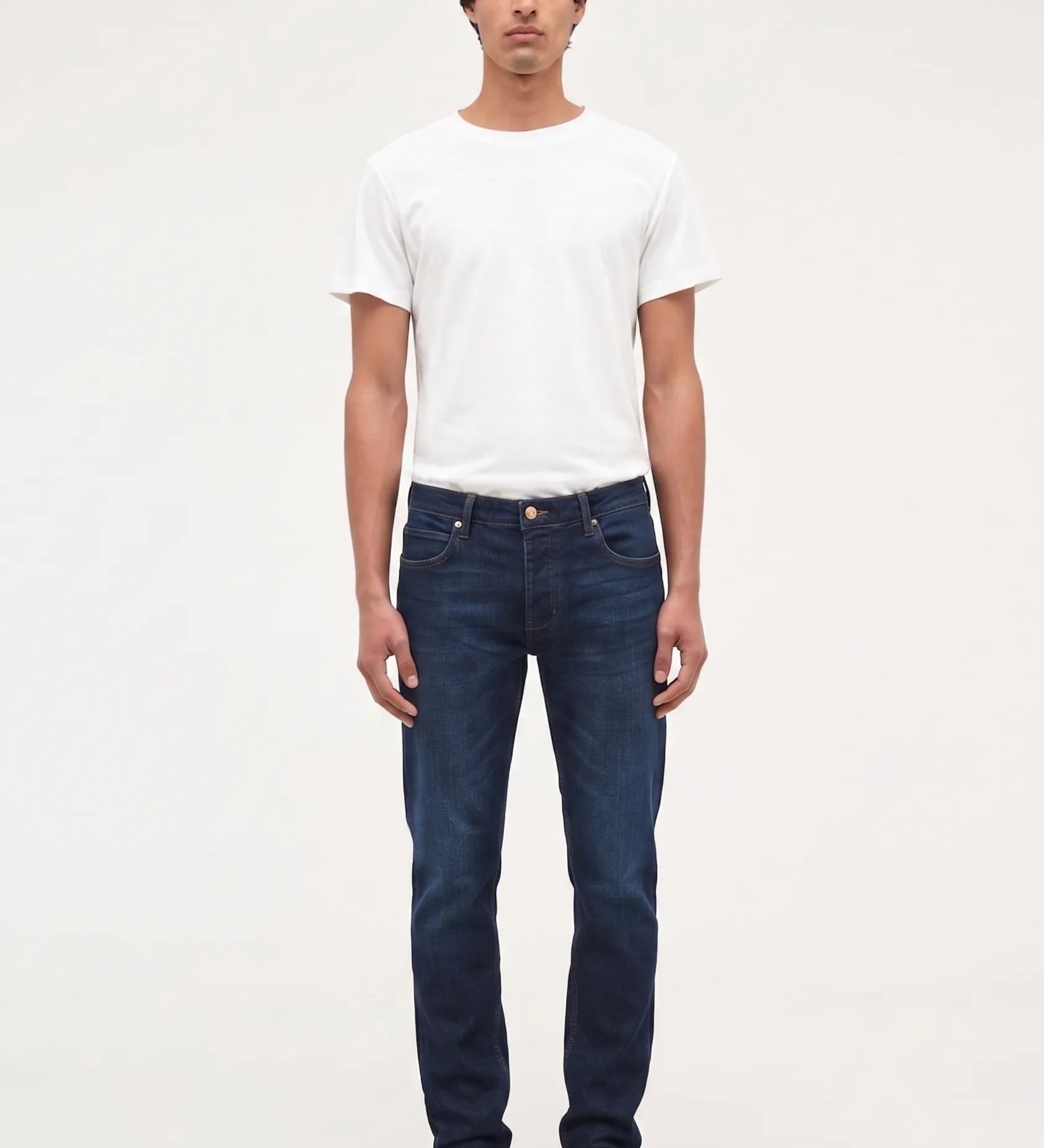 A man wearing dark blue slim-fit jeans, a white t-shirt, and black shoes, standing against a plain white studio background.
