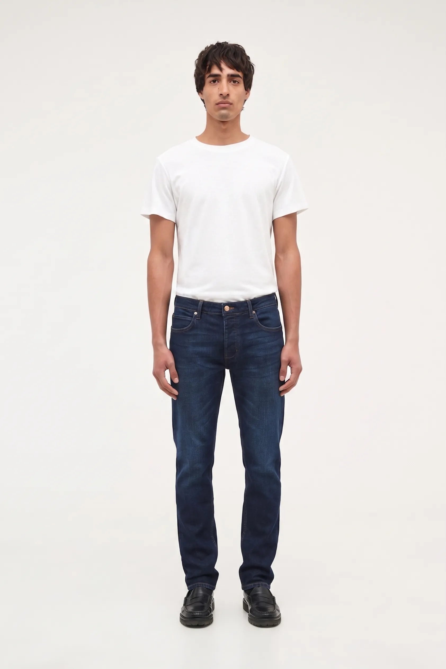 A man wearing dark blue slim-fit jeans, a white t-shirt, and black shoes, standing against a plain white studio background.