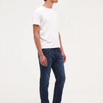 A man standing in profile wearing a plain white short-sleeve t-shirt, dark blue slim-fit jeans, and black leather shoes against a solid white studio background.