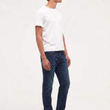 A man standing in profile wearing a plain white short-sleeve t-shirt, dark blue slim-fit jeans, and black leather shoes against a solid white studio background.