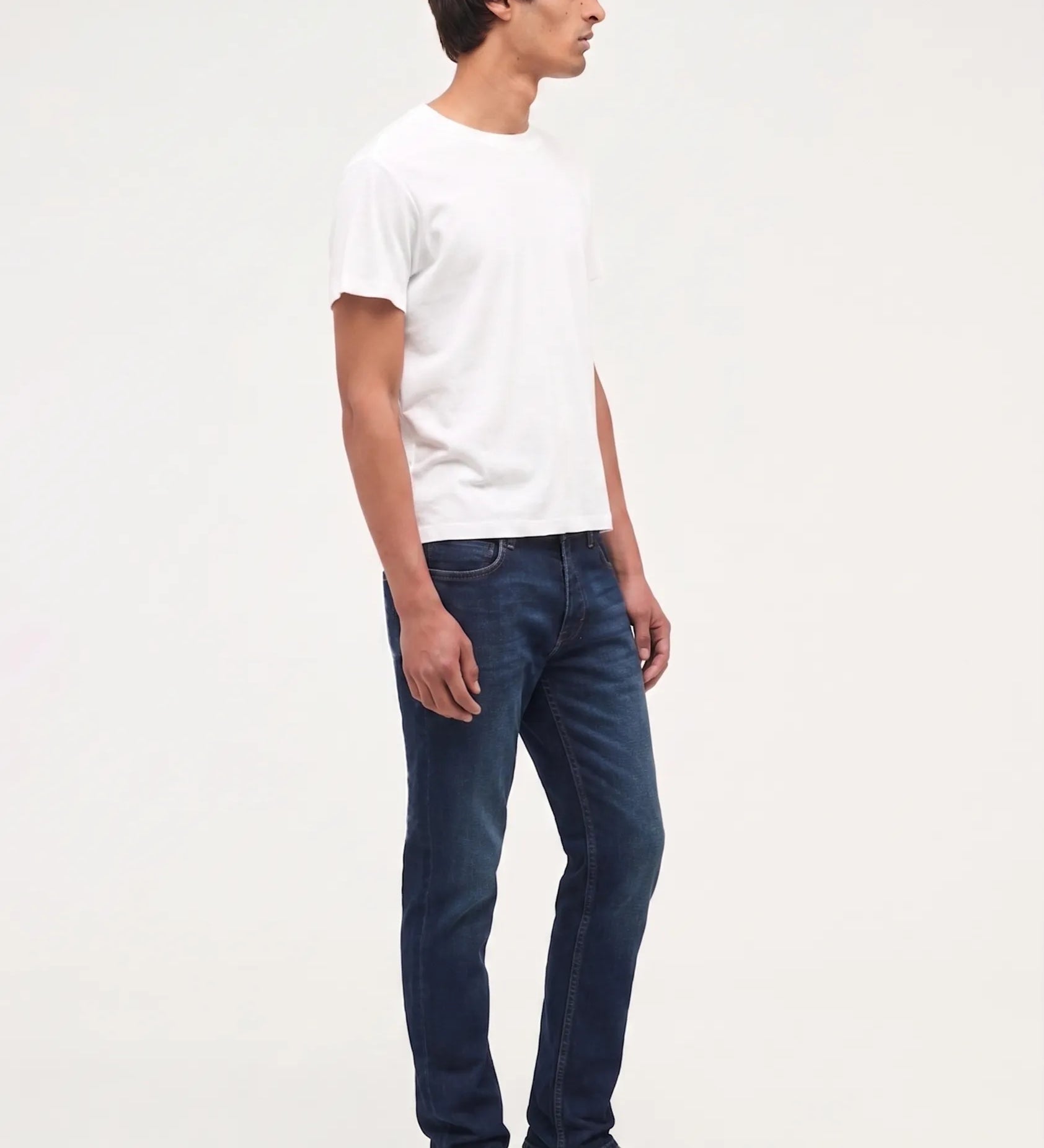 A man standing in profile wearing a plain white short-sleeve t-shirt, dark blue slim-fit jeans, and black leather shoes against a solid white studio background.