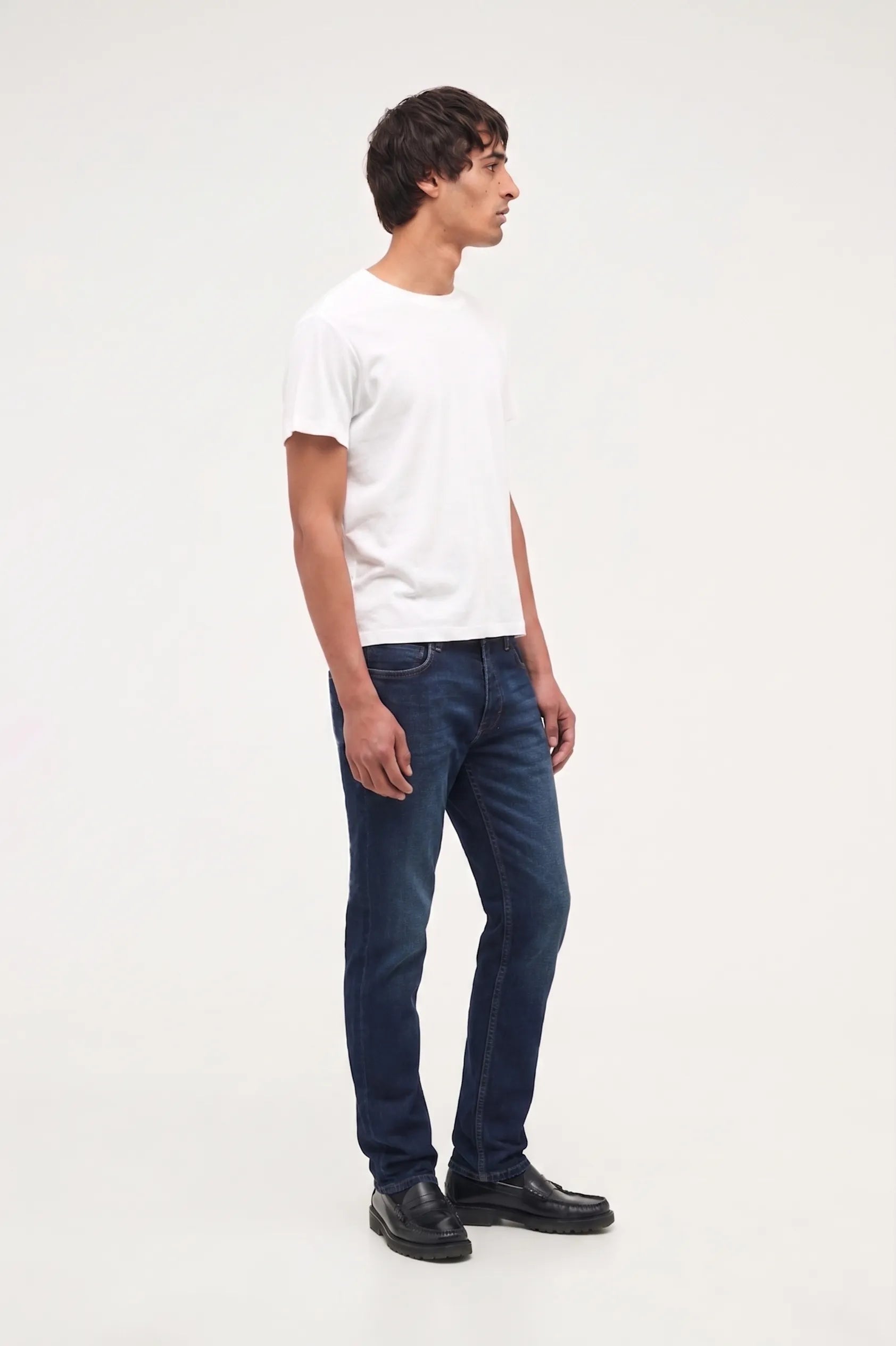 A man standing in profile wearing a plain white short-sleeve t-shirt, dark blue slim-fit jeans, and black leather shoes against a solid white studio background.