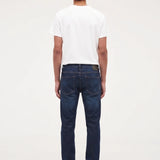 A man viewed from behind wearing a white t-shirt, dark blue jeans, and black shoes, standing against a plain white studio background.