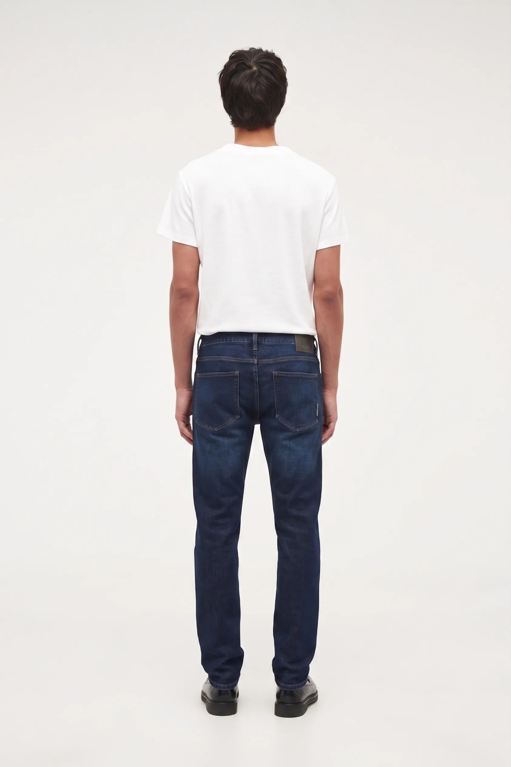 A man viewed from behind wearing a white t-shirt, dark blue jeans, and black shoes, standing against a plain white studio background.
