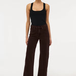 A model stands against a plain white studio background wearing dark brown wide-leg corduroy pants, a black tank top, and black pointed shoes.