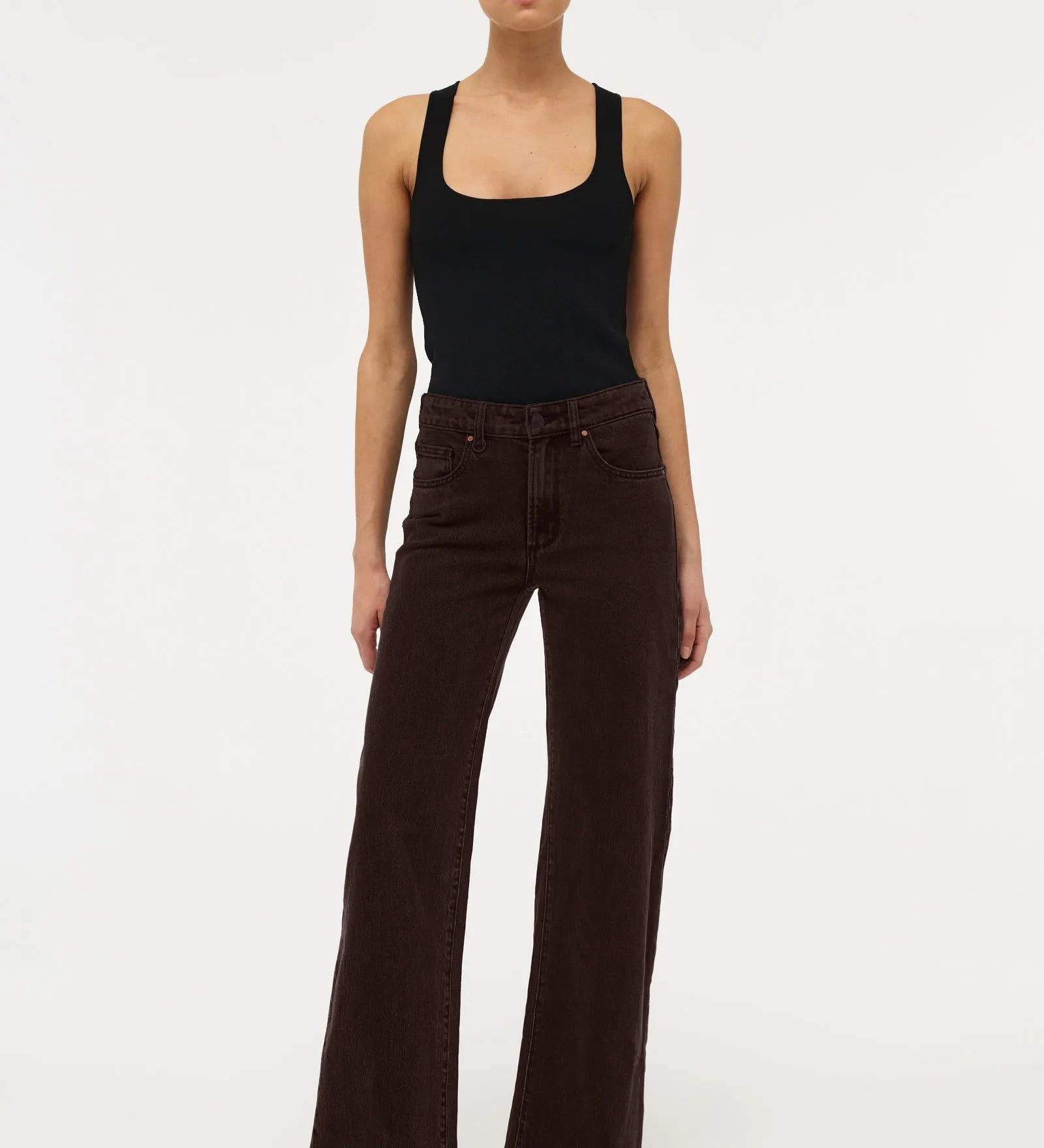 A model stands against a plain white studio background wearing dark brown wide-leg corduroy pants, a black tank top, and black pointed shoes.