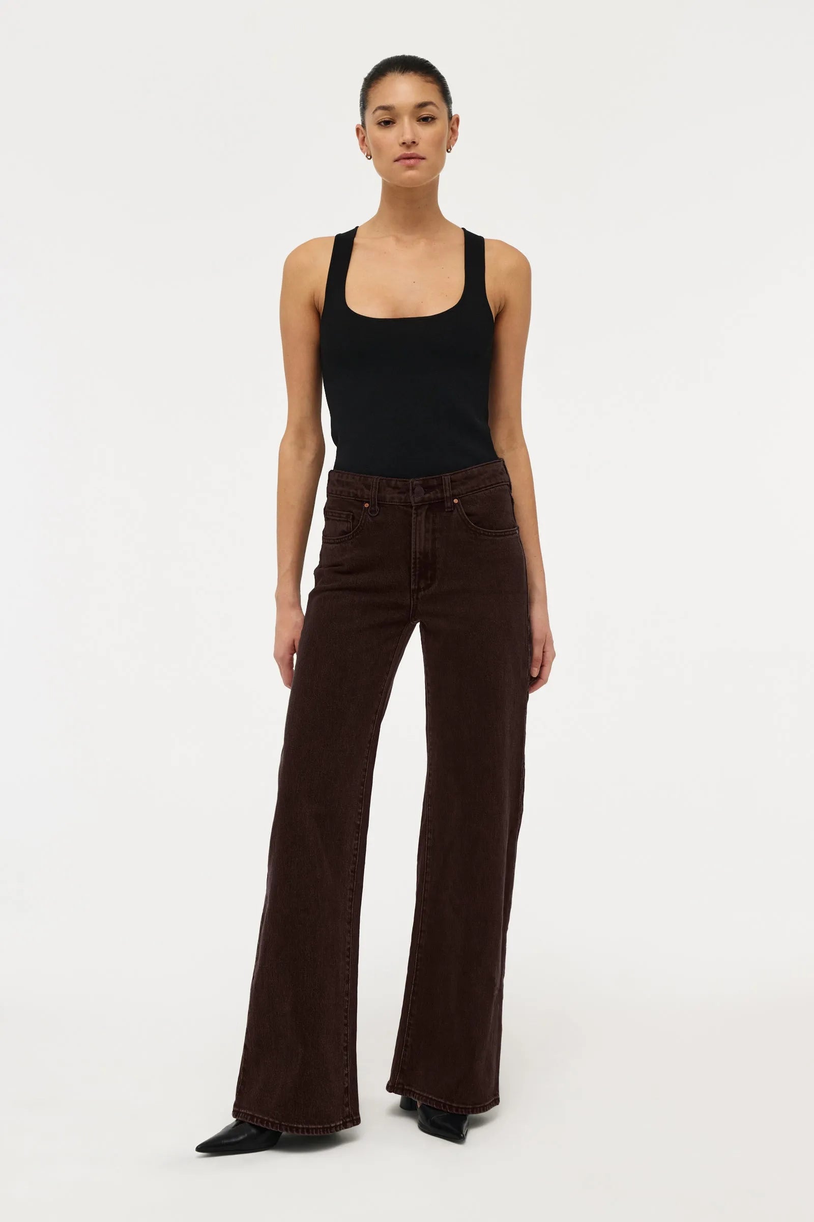 A model stands against a plain white studio background wearing dark brown wide-leg corduroy pants, a black tank top, and black pointed shoes.