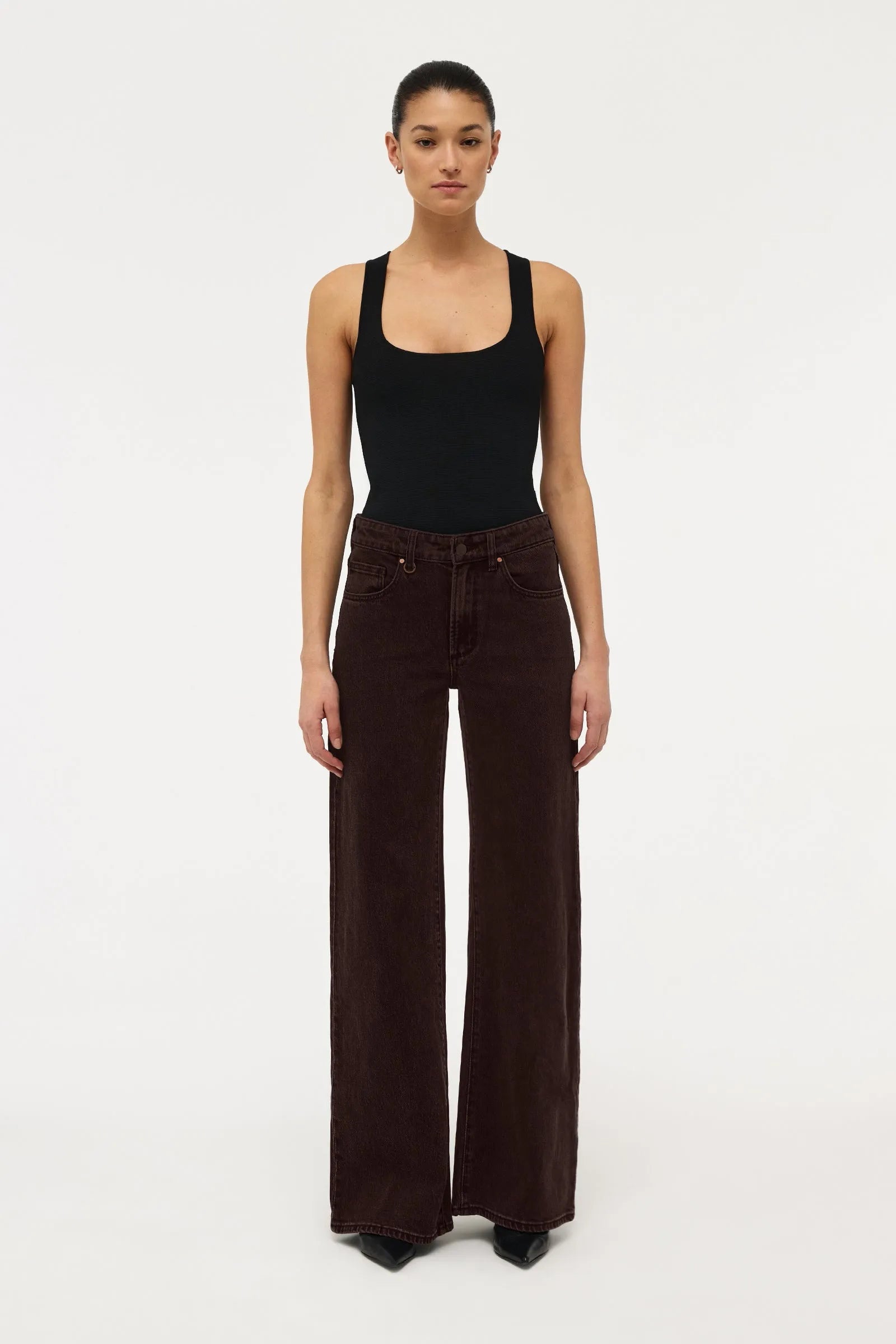 A model stands against a plain white studio background wearing a black sleeveless top and dark brown wide-leg corduroy pants.
