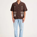A man modeling a brown short-sleeve button-up shirt with white and multicolor embroidered patterns on the front, paired with light blue jeans and gray sneakers, against a plain off-white studio background.