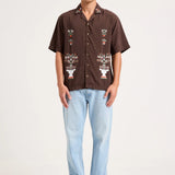 A man modeling a brown short-sleeve button-up shirt with white and multicolor embroidered patterns on the front, paired with light blue jeans and gray sneakers, against a plain off-white studio background.