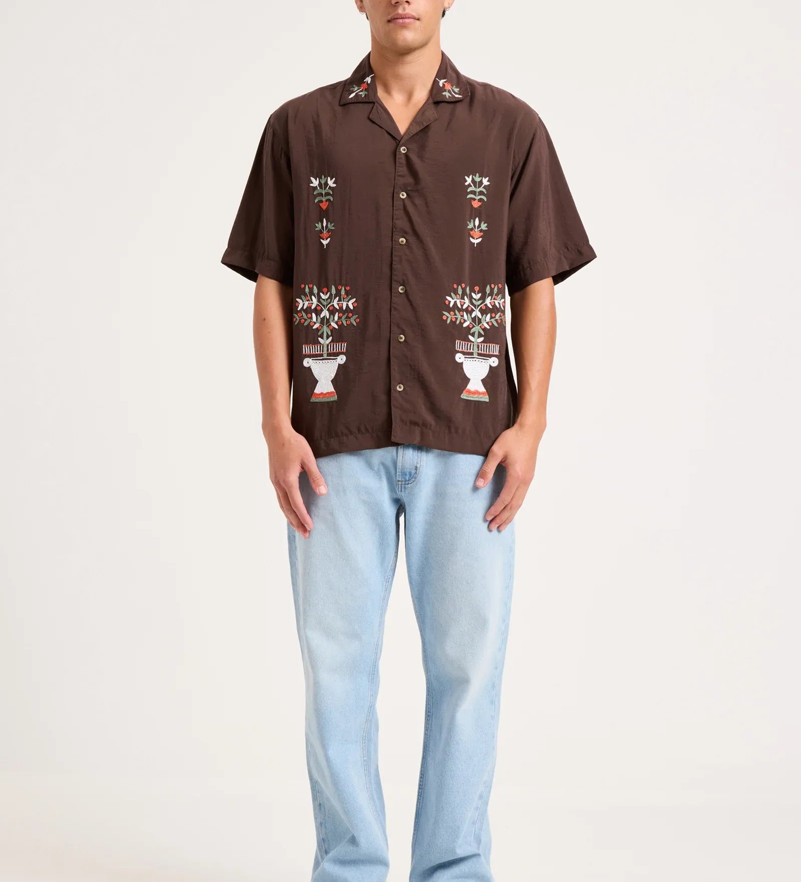 A man modeling a brown short-sleeve button-up shirt with white and multicolor embroidered patterns on the front, paired with light blue jeans and gray sneakers, against a plain off-white studio background.