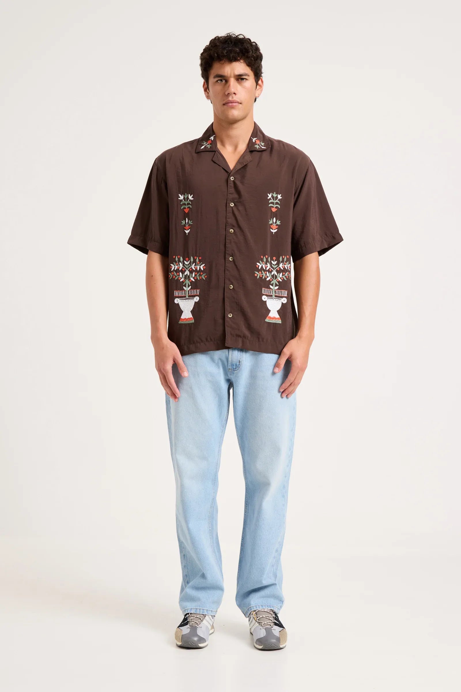 A man modeling a brown short-sleeve button-up shirt with white and multicolor embroidered patterns on the front, paired with light blue jeans and gray sneakers, against a plain off-white studio background.