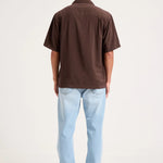 A man viewed from behind wearing a dark brown short-sleeve button-up shirt and light blue jeans, standing against a plain white studio background.