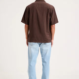 A man viewed from behind wearing a dark brown short-sleeve button-up shirt and light blue jeans, standing against a plain white studio background.