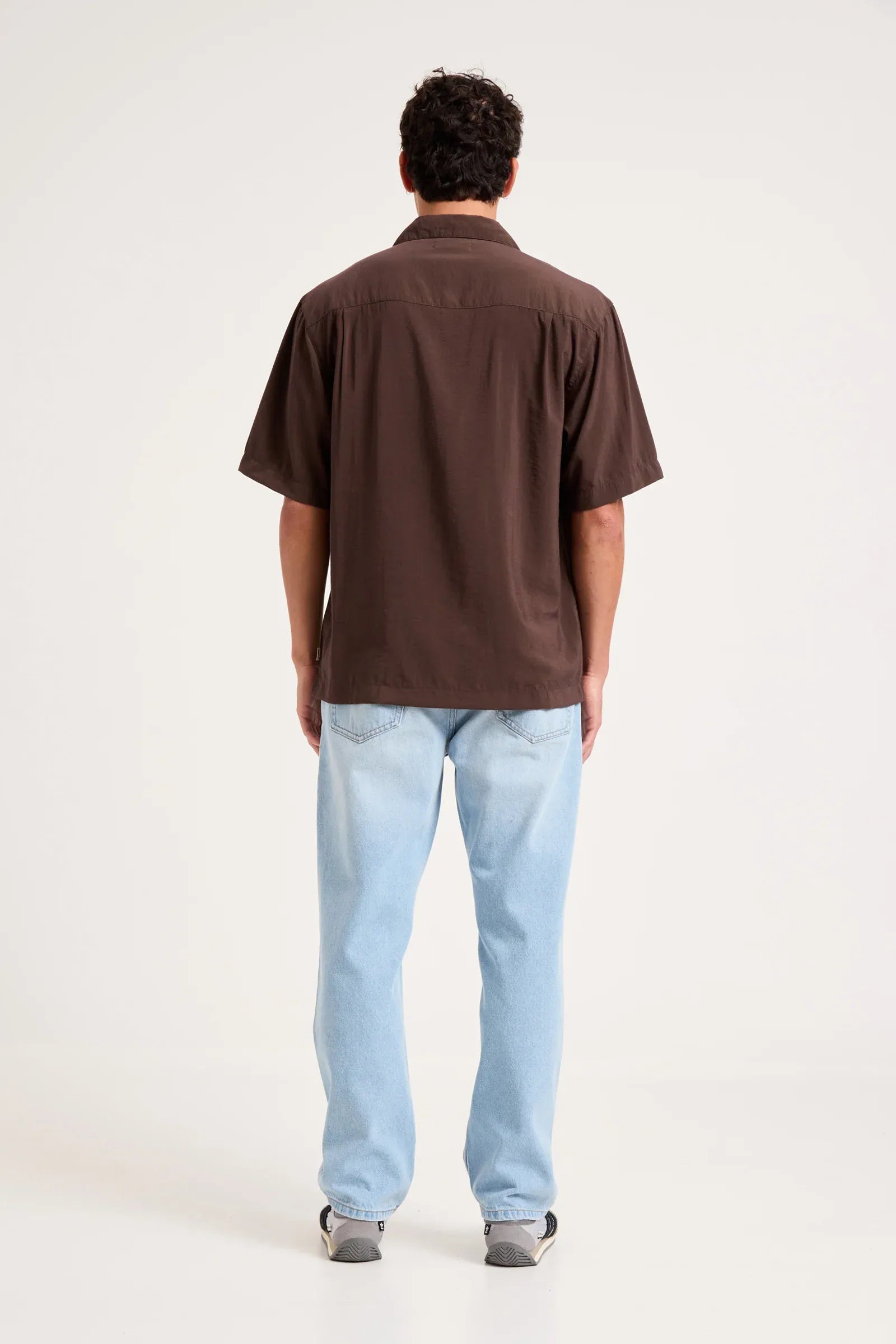 A man viewed from behind wearing a dark brown short-sleeve button-up shirt and light blue jeans, standing against a plain white studio background.