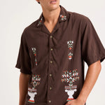 A man wearing a brown short-sleeve button-up shirt with colorful embroidered floral and geometric patterns, paired with light blue jeans, photographed against a plain light background.