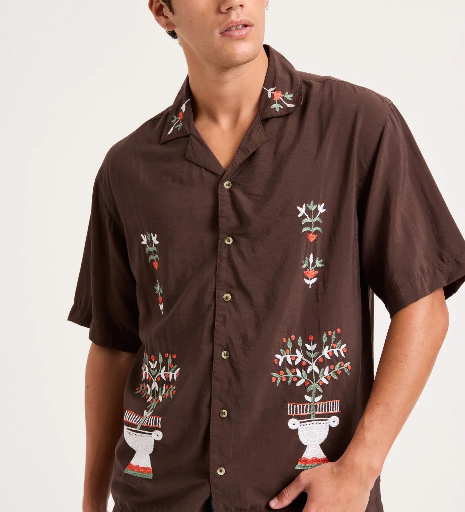 A man wearing a brown short-sleeve button-up shirt with colorful embroidered floral and geometric patterns, paired with light blue jeans, photographed against a plain light background.
