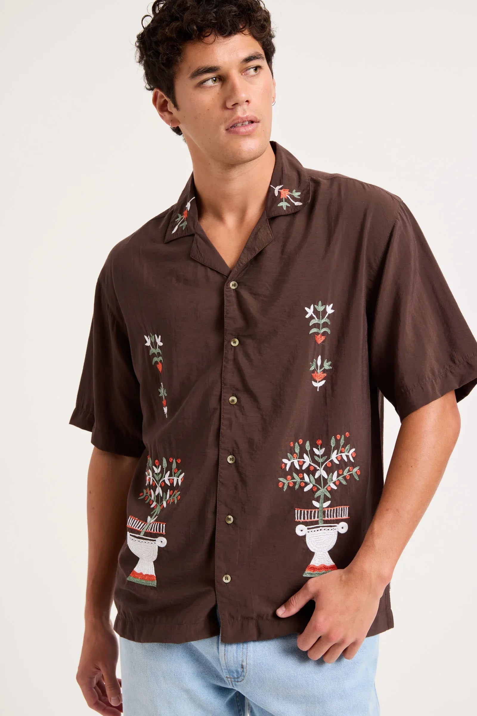 A man wearing a brown short-sleeve button-up shirt with colorful embroidered floral and geometric patterns, paired with light blue jeans, photographed against a plain light background.