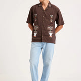 A man modeling a brown short-sleeve button-up shirt with colorful floral embroidery on the front, paired with light blue jeans and gray sneakers, against a plain white studio background.