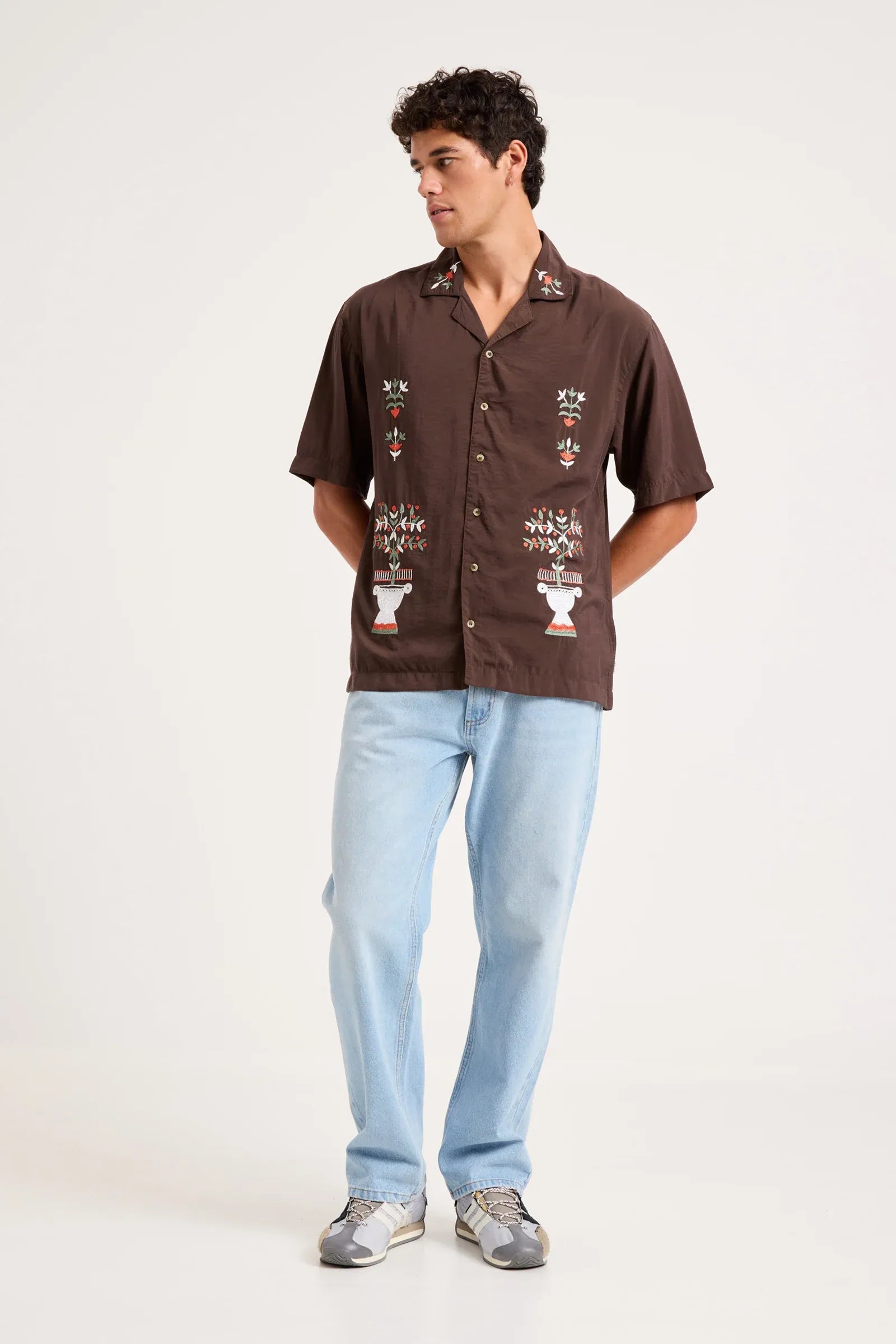 A man modeling a brown short-sleeve button-up shirt with colorful floral embroidery on the front, paired with light blue jeans and gray sneakers, against a plain white studio background.