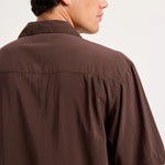A man wearing a dark brown short-sleeve button-up shirt, shown from a side-back angle against a plain light background.