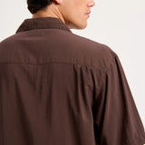 A man wearing a dark brown short-sleeve button-up shirt, shown from a side-back angle against a plain light background.