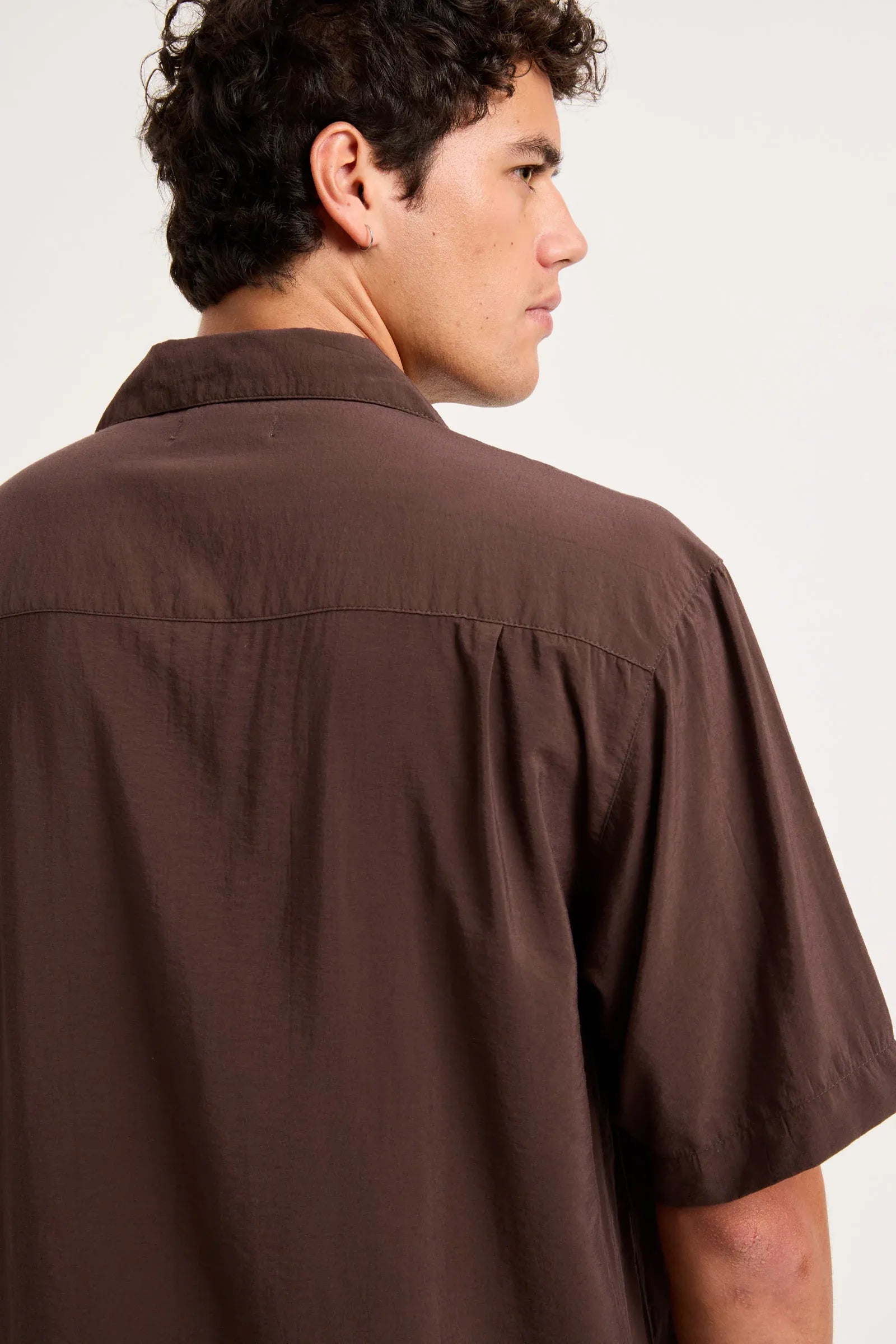 A man wearing a dark brown short-sleeve button-up shirt, shown from a side-back angle against a plain light background.