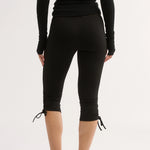 Rear view of a model wearing black long-sleeve capri leggings with tie-up details at the hem, paired with a black long-sleeve top and black mule heels, against a plain white studio background.