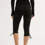 Rear view of a model wearing black long-sleeve capri leggings with tie-up details at the hem, paired with a black long-sleeve top and black mule heels, against a plain white studio background.