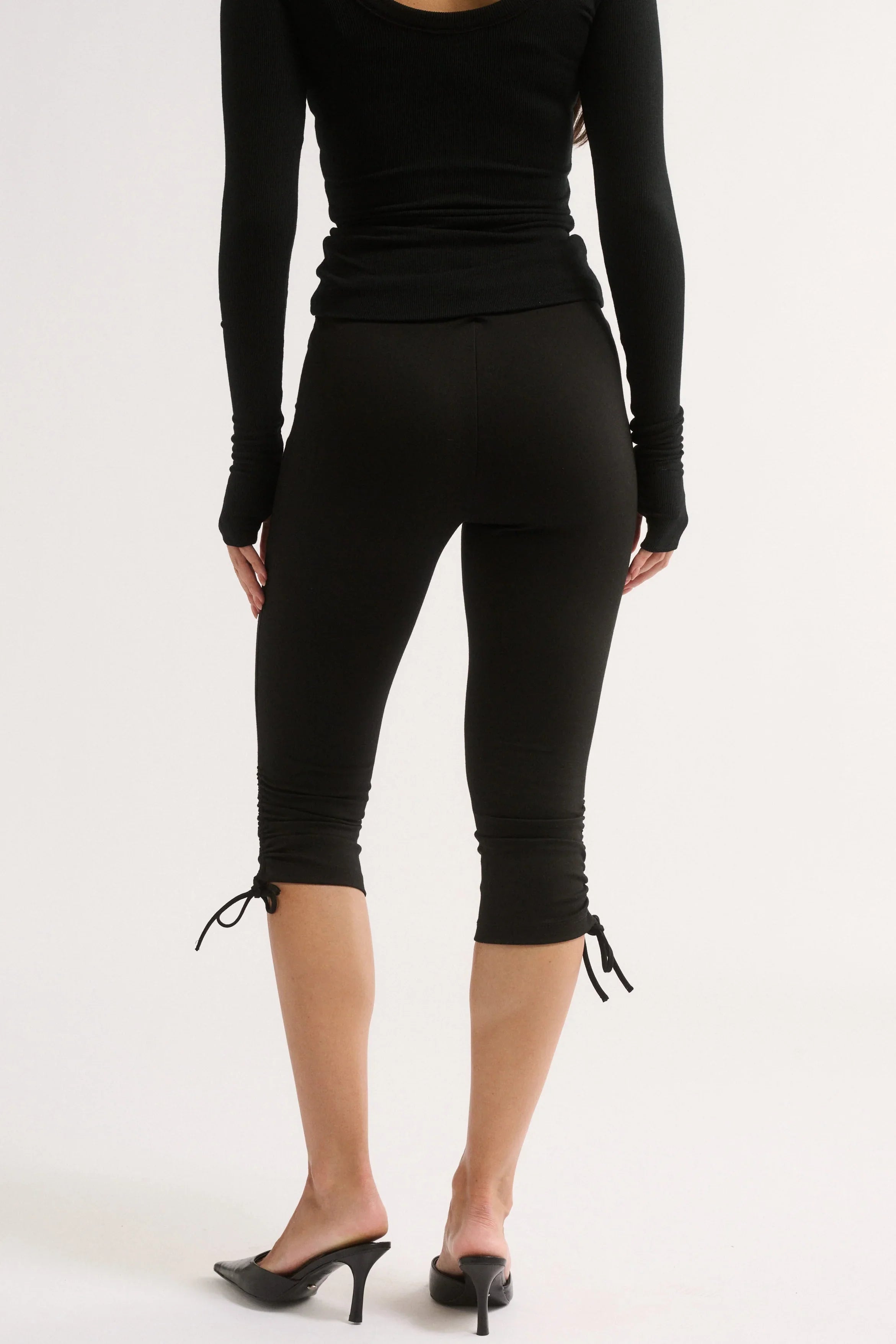 Rear view of a model wearing black long-sleeve capri leggings with tie-up details at the hem, paired with a black long-sleeve top and black mule heels, against a plain white studio background.