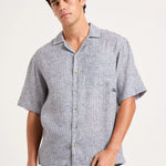 A man wearing a short-sleeve button-up camp shirt with a light blue and white micro-pattern, paired with brown pants, photographed against a solid off-white background.