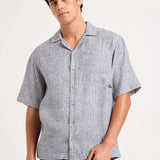 A man wearing a short-sleeve button-up camp shirt with a light blue and white micro-pattern, paired with brown pants, photographed against a solid off-white background.