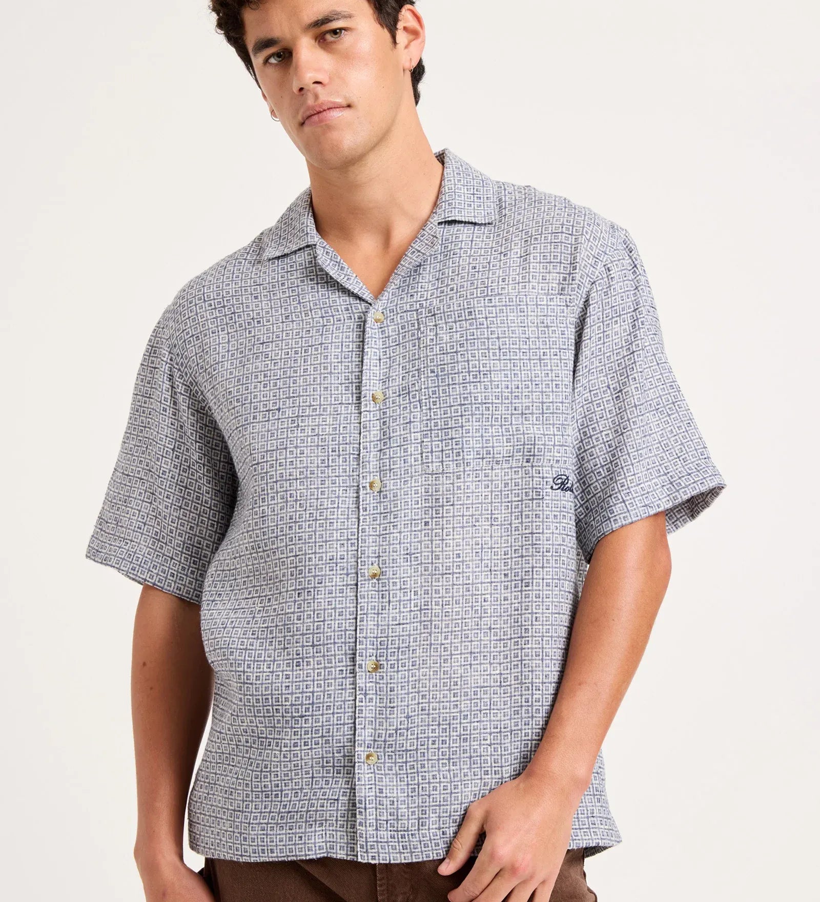 A man wearing a short-sleeve button-up camp shirt with a light blue and white micro-pattern, paired with brown pants, photographed against a solid off-white background.
