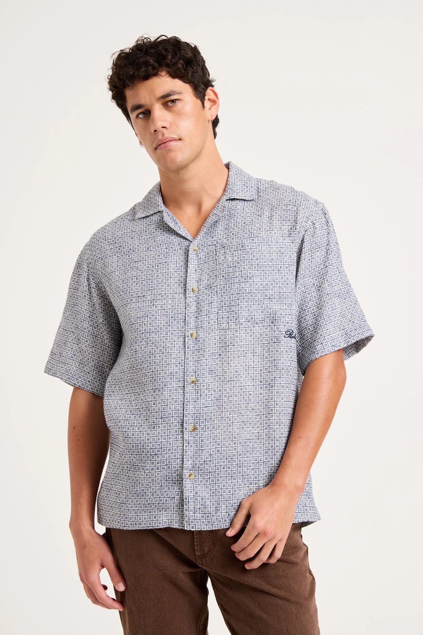 A man wearing a short-sleeve button-up camp shirt with a light blue and white micro-pattern, paired with brown pants, photographed against a solid off-white background.