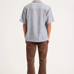 A man viewed from behind wearing a light gray short-sleeve button-up shirt with a subtle textured pattern, paired with brown pants and gray sneakers, against a plain white studio background.