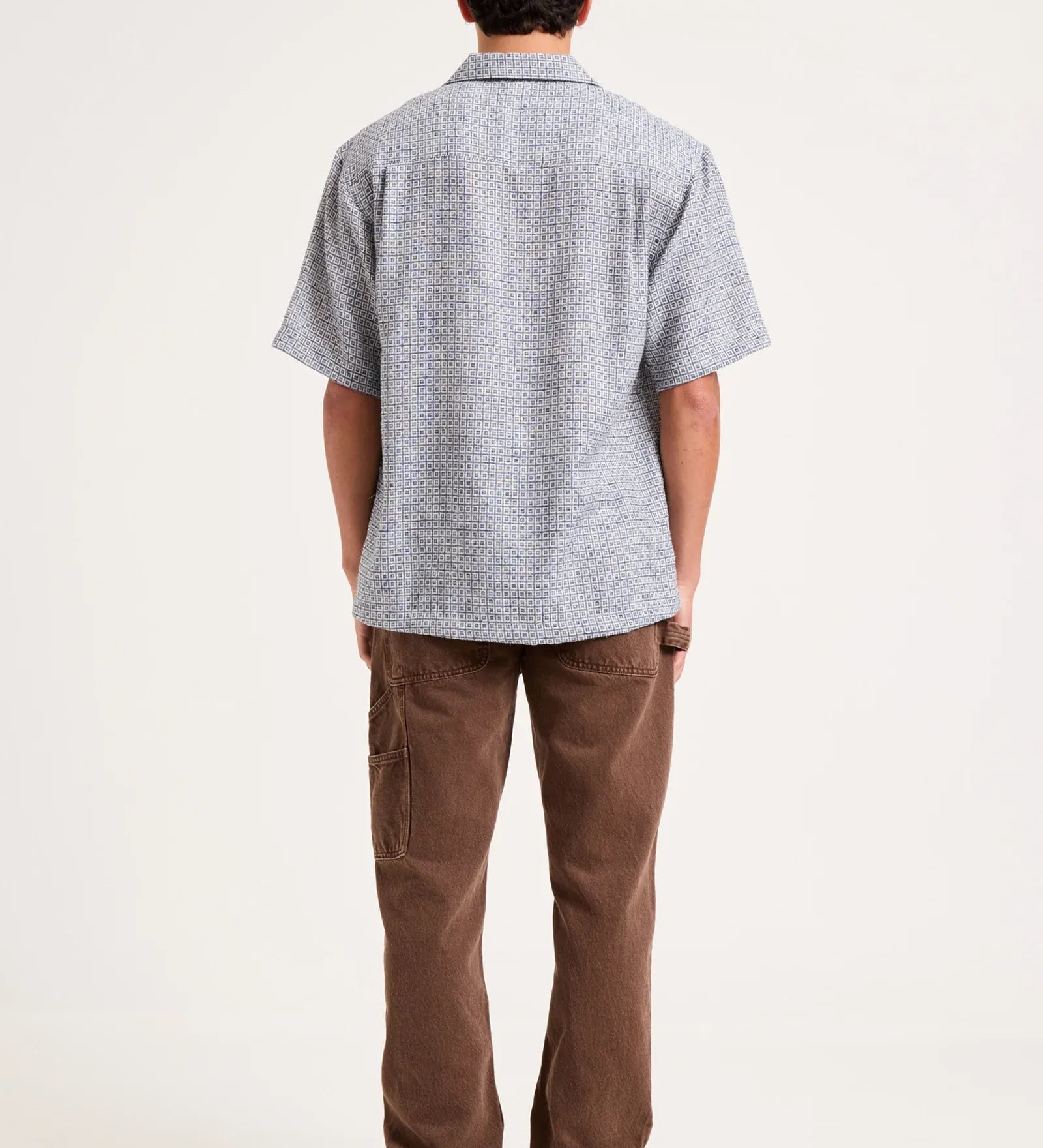 A man viewed from behind wearing a light gray short-sleeve button-up shirt with a subtle textured pattern, paired with brown pants and gray sneakers, against a plain white studio background.
