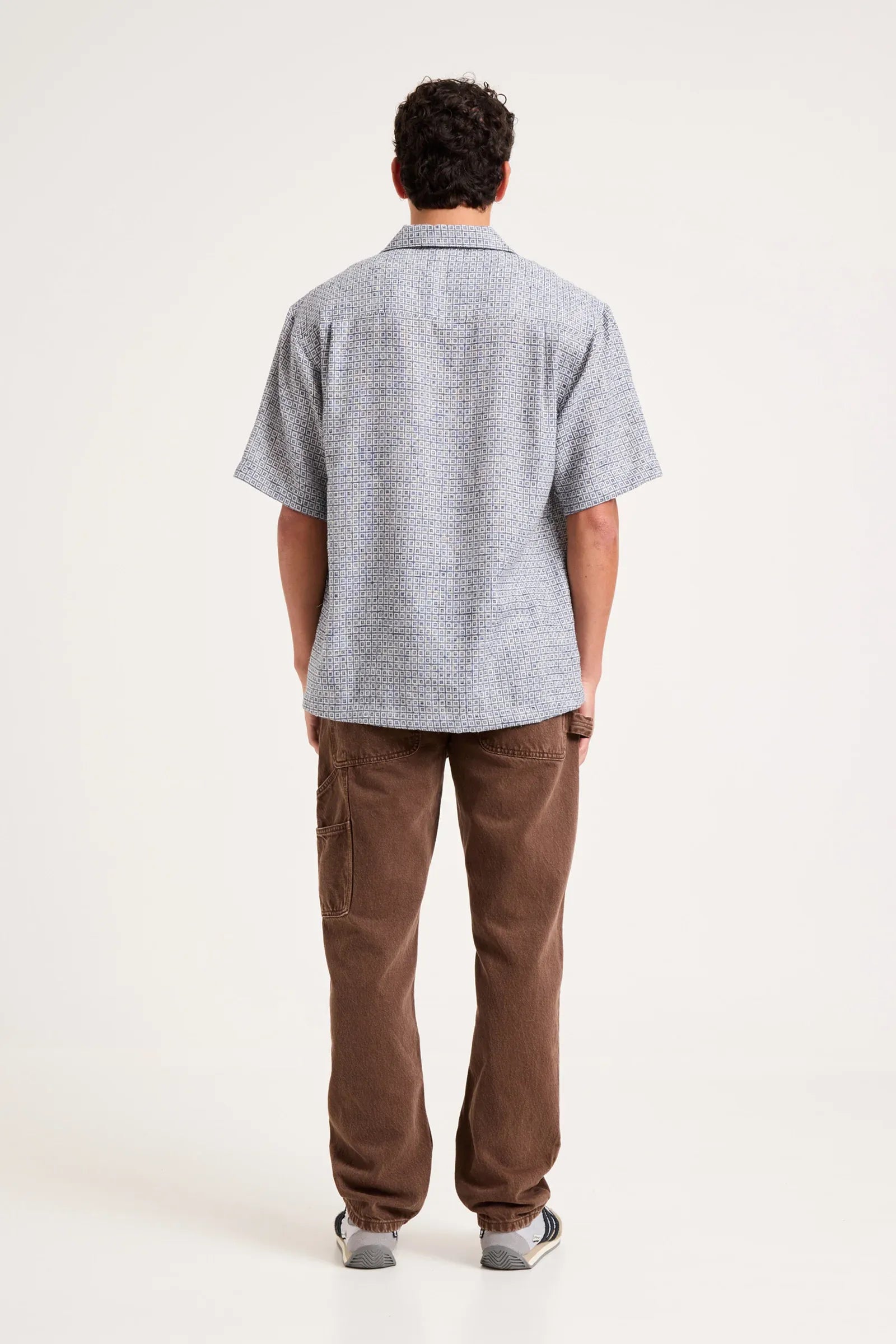 A man viewed from behind wearing a light gray short-sleeve button-up shirt with a subtle textured pattern, paired with brown pants and gray sneakers, against a plain white studio background.
