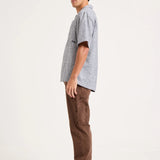 A man standing in profile wearing a light gray short-sleeve t-shirt, brown cargo pants, and gray sneakers with black stripes, photographed against a plain off-white studio background.