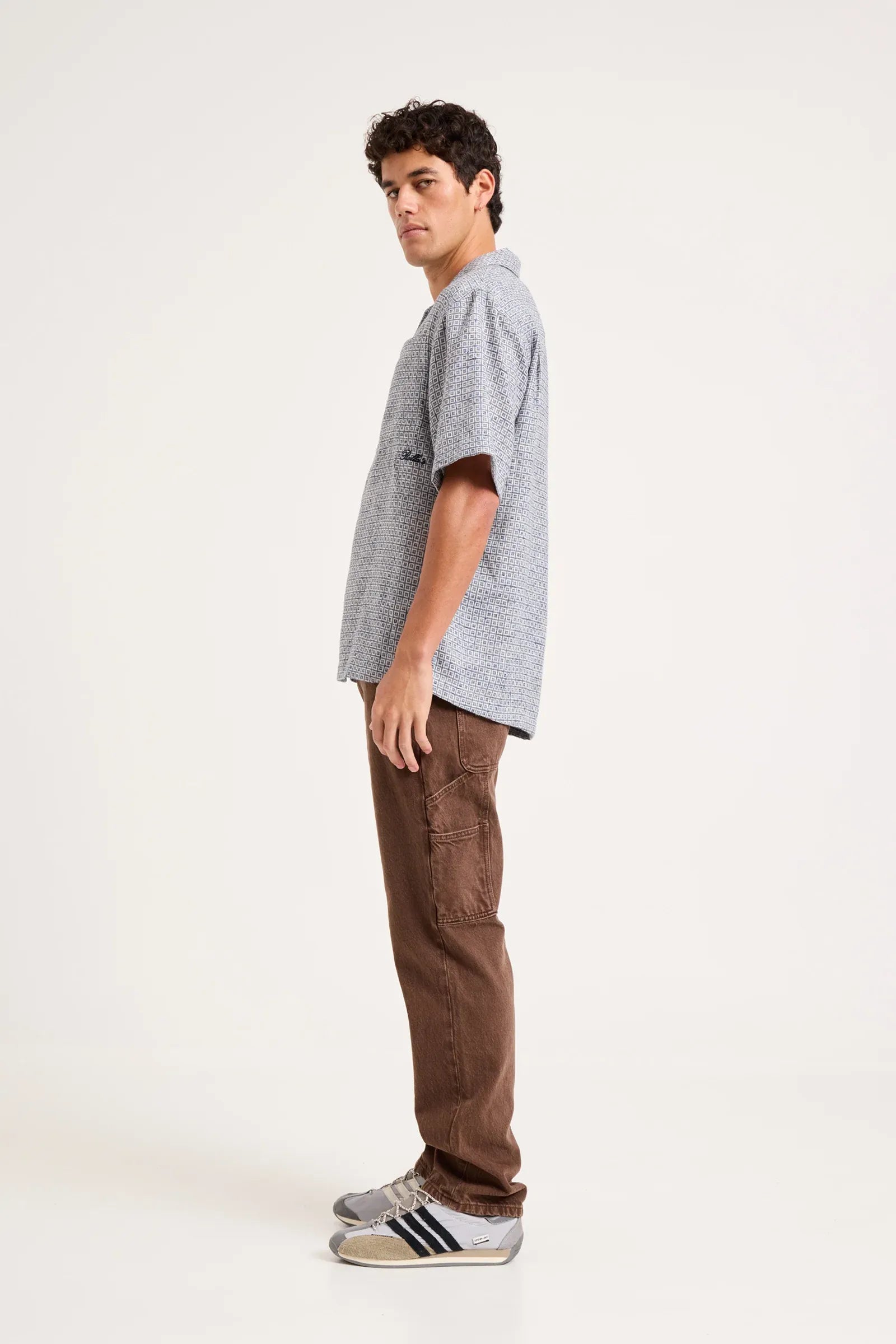 A man standing in profile wearing a light gray short-sleeve t-shirt, brown cargo pants, and gray sneakers with black stripes, photographed against a plain off-white studio background.