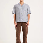 A man modeling a light gray short-sleeve button-up shirt and brown pants against a plain off-white studio background.