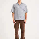 A man modeling a light gray short-sleeve button-up shirt and brown pants against a plain off-white studio background.