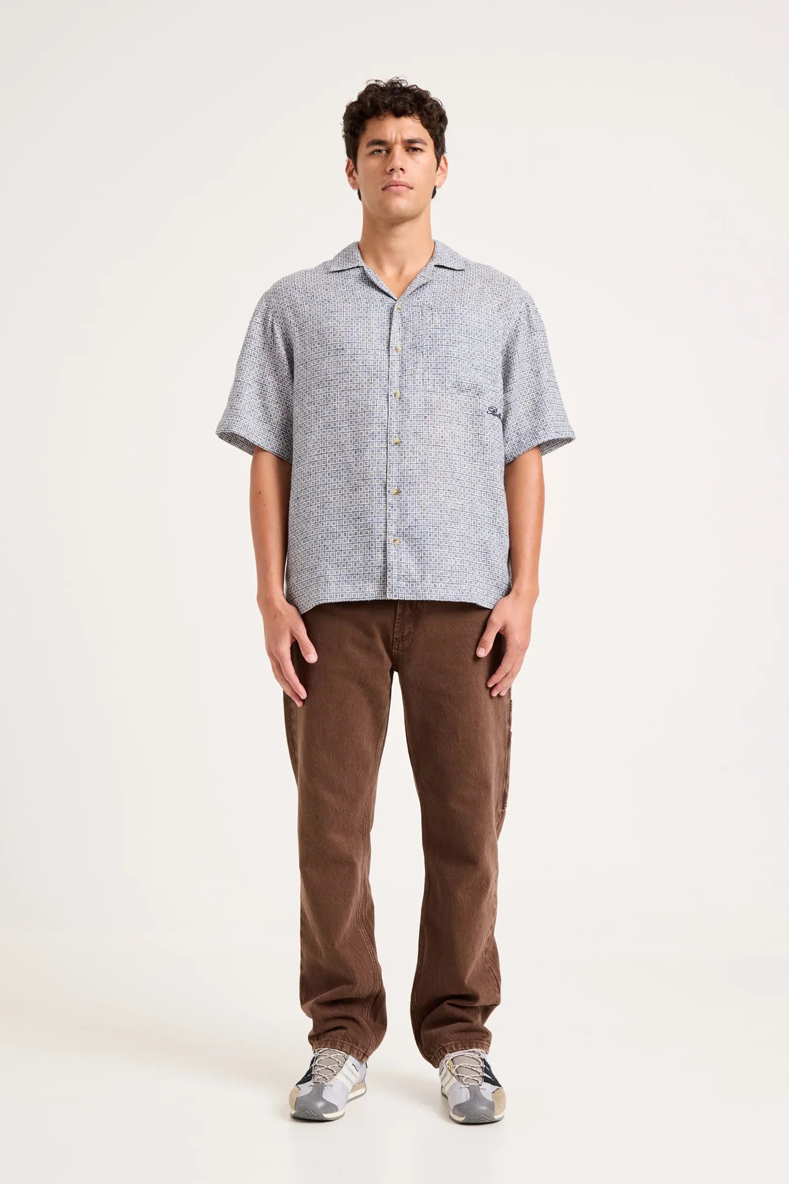 A man modeling a light gray short-sleeve button-up shirt and brown pants against a plain off-white studio background.