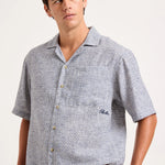 A man wearing a short-sleeve button-up camp shirt with a light blue and white micro-pattern, featuring a chest pocket with a small embroidered logo.