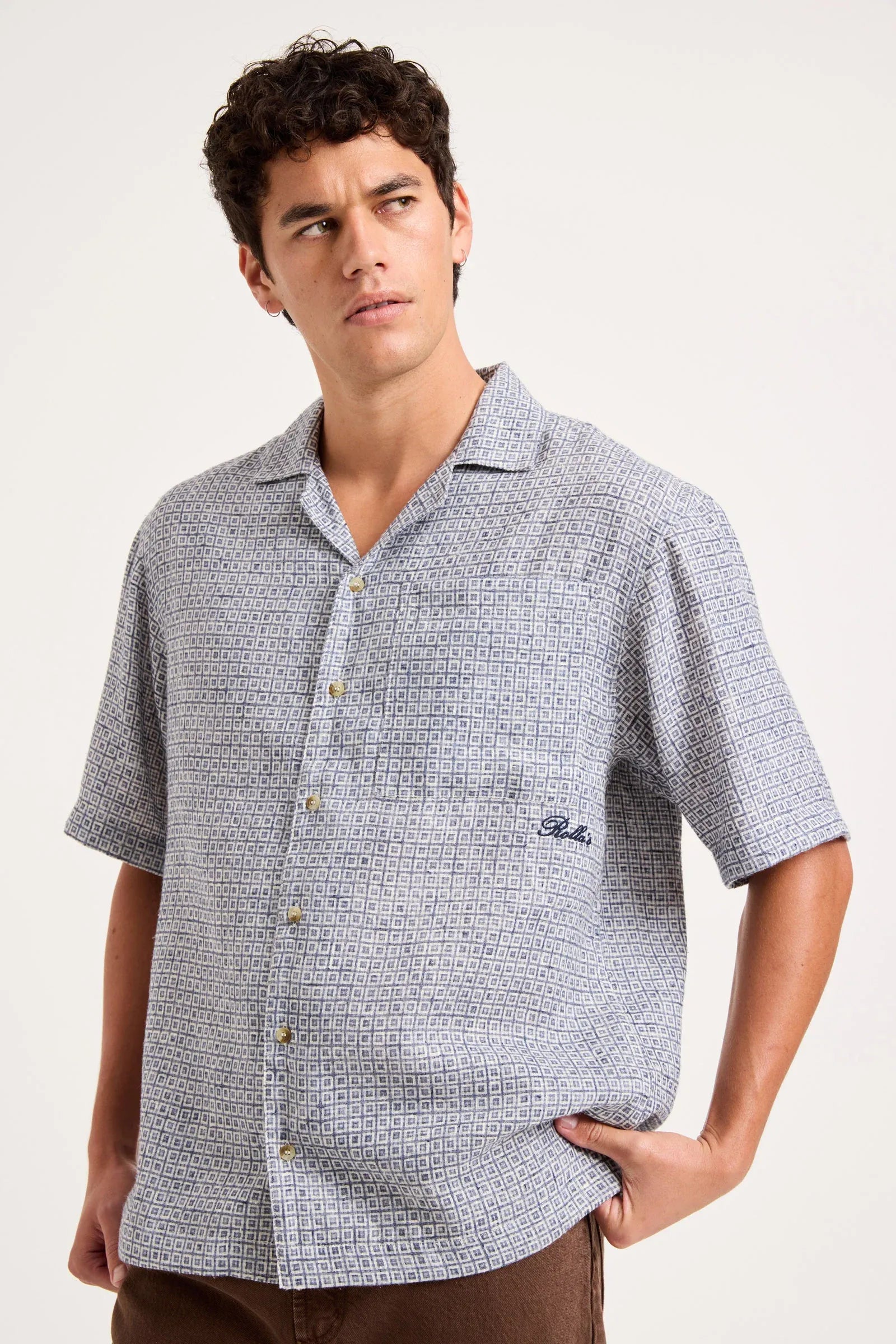 A man wearing a short-sleeve button-up camp shirt with a light blue and white micro-pattern, featuring a chest pocket with a small embroidered logo.