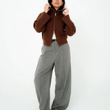 A woman stands against a plain white studio background wearing a cropped brown ribbed zip-front jacket, wide-leg gray plaid trousers, a white top, and black pointed shoes.