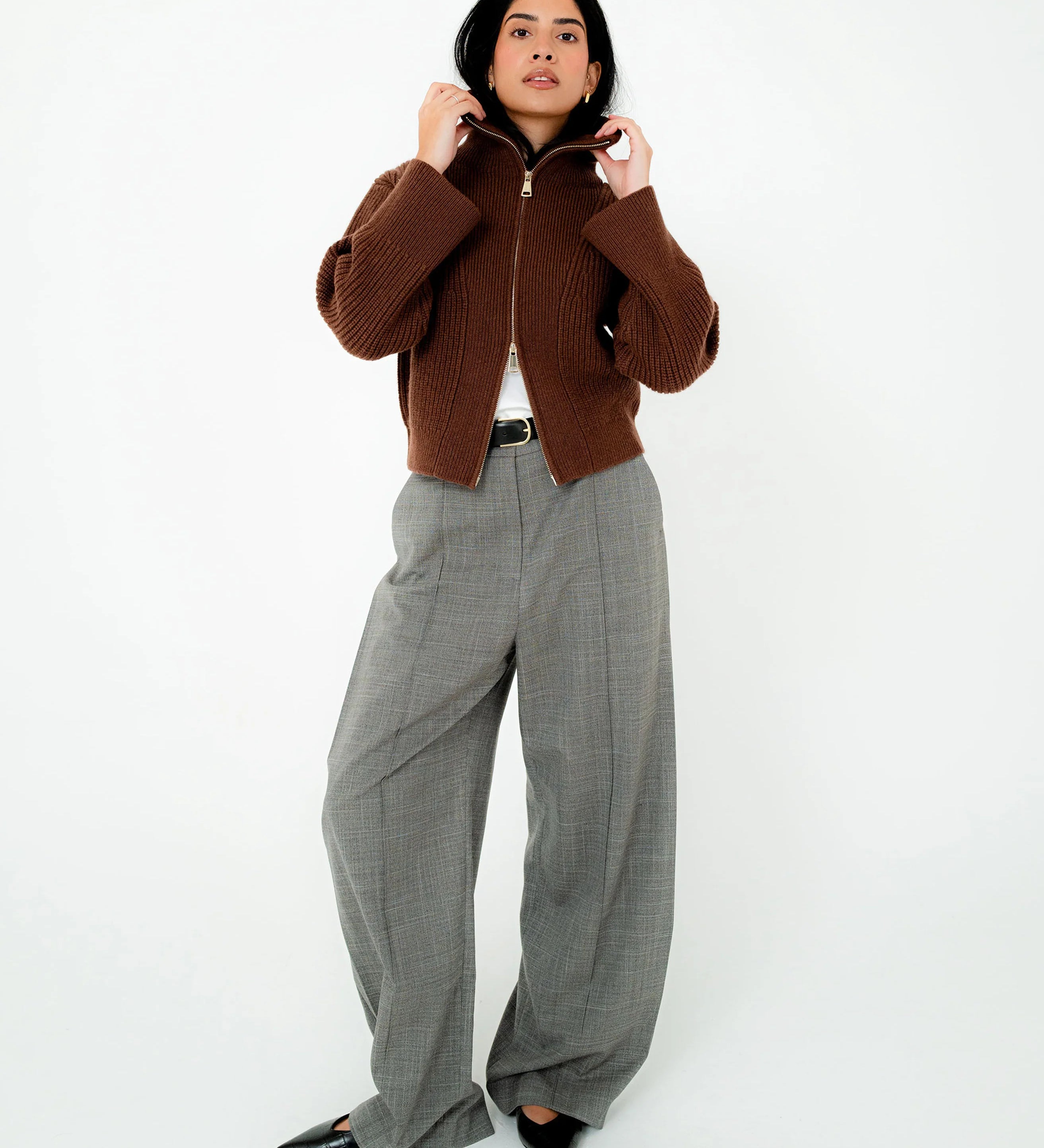 A woman stands against a plain white studio background wearing a cropped brown ribbed zip-front jacket, wide-leg gray plaid trousers, a white top, and black pointed shoes.
