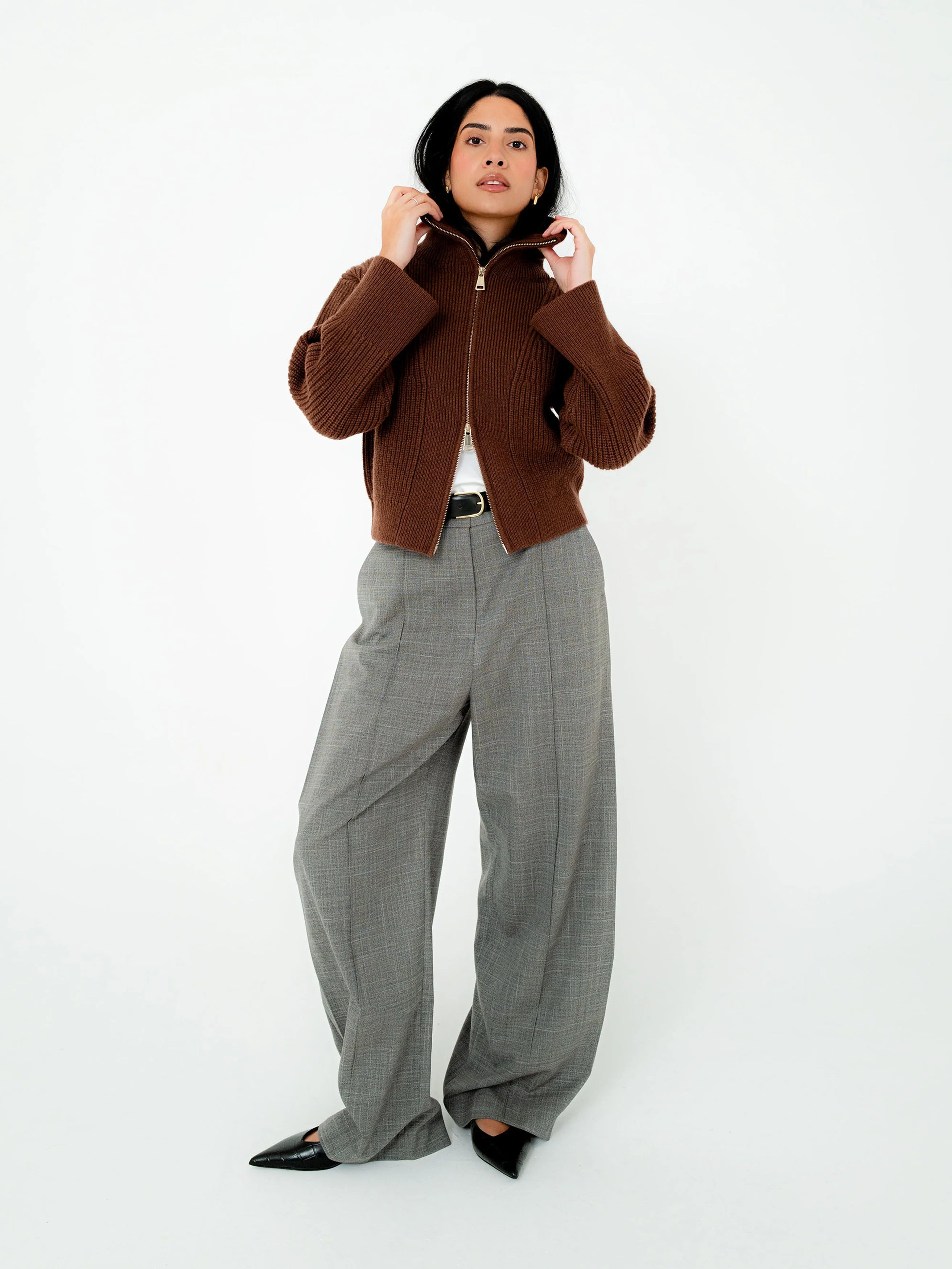 A woman stands against a plain white studio background wearing a cropped brown ribbed zip-front jacket, wide-leg gray plaid trousers, a white top, and black pointed shoes.