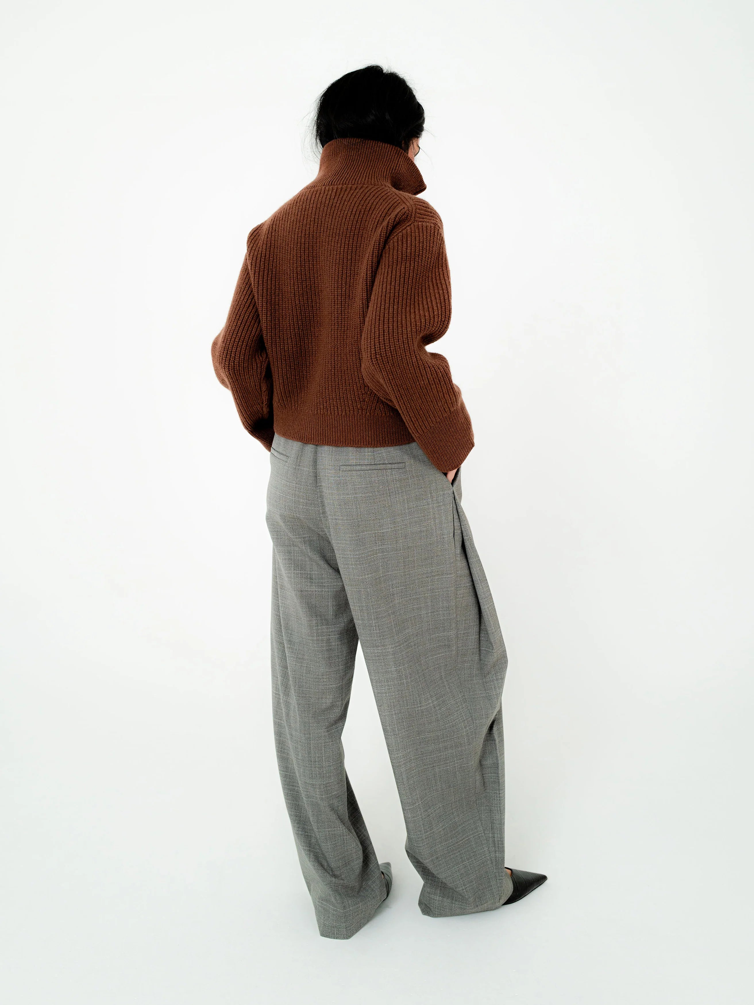 A person shown from the back wearing a brown ribbed knit mock-neck sweater and wide-leg gray trousers, standing against a plain white studio background.