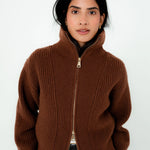 A woman wearing a brown ribbed knit zip-up sweater with a high stand-up collar, paired with gray trousers, against a plain white background.